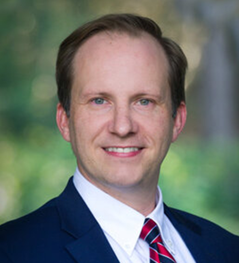 A professional headshot of a Caucasian man with short, light brown hair and a friendly smile. He is wearing a navy blue suit, a white dress shirt, and a red and blue striped tie. The background is blurred with lush green foliage, suggesting an outdoor setting with natural lighting.