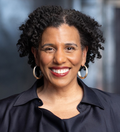 A professional headshot of a smiling Black woman with short, naturally curly hair. She is wearing a navy blue button-up shirt with an open collar and large gold hoop earrings. Her warm expression conveys confidence and approachability. The blurred background suggests an office window with soft, natural lighting.