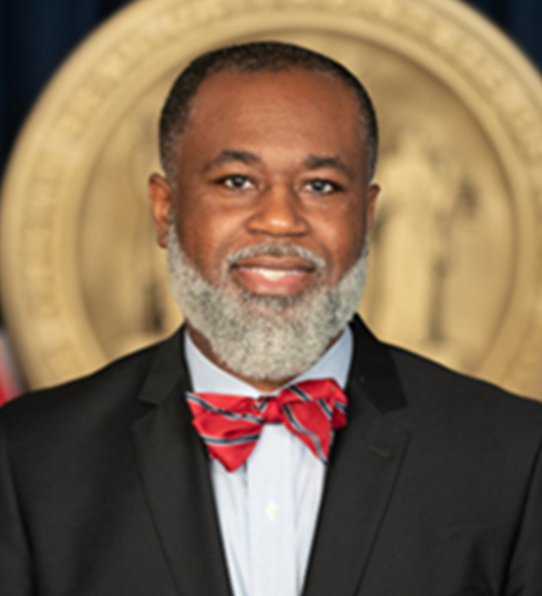 A professional headshot of a Black man with a neatly trimmed gray beard and short hair. He is wearing a black suit, light blue dress shirt, and a red bow tie with a subtle pattern. He has a warm smile and a confident expression. The background features a large gold seal, slightly blurred, suggesting an official or governmental setting.