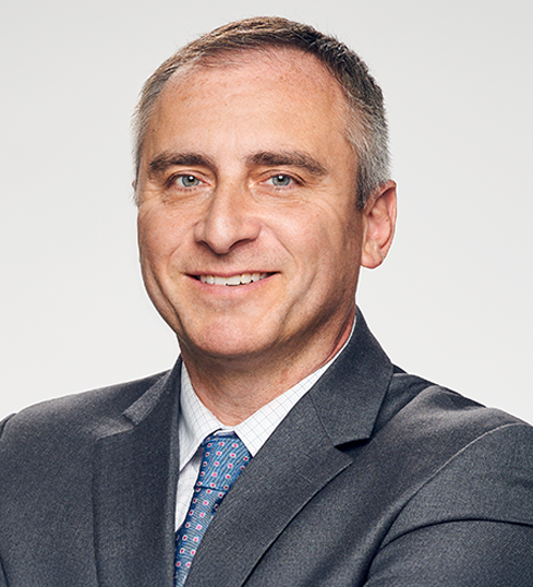 Professional portrait of a male in business attire. He is smiling. He is wearing a grey suit and white dress shirt with a light blue tie featuring a red dotted pattern.