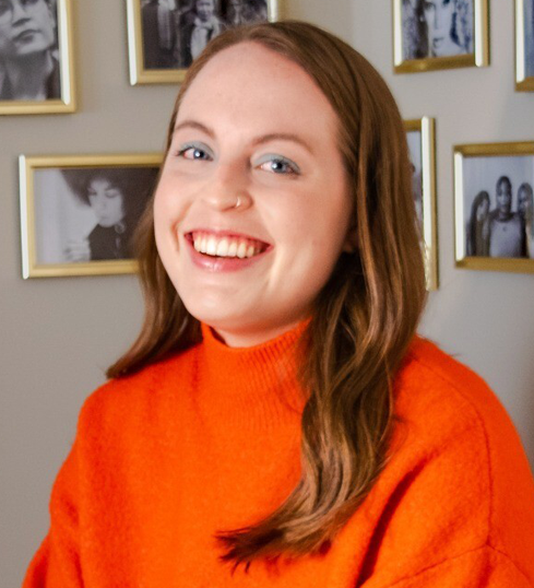 A young woman with fair skin and long, wavy brown hair smiles warmly at the camera. She wears a vibrant orange turtleneck sweater. Her blue eyeshadow and small nose ring add to her distinctive look. Behind her, a wall is adorned with framed black-and-white photographs, arranged neatly in gold frames. The soft lighting and her joyful expression create a welcoming and friendly atmosphere.